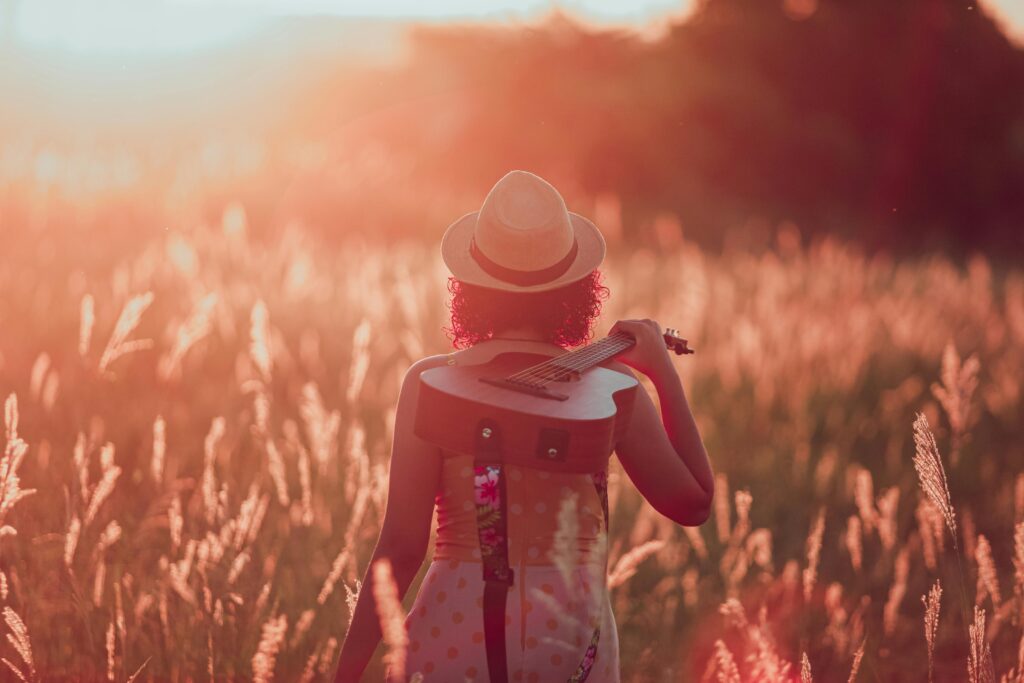 A woman in a hat carrying a guitar through a sunlit field at sunset in Paragominas, Brazil.