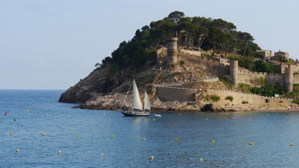 Scenic view of Tossa de Mar's historic castle overlooking the Mediterranean with a sailboat in the foreground.